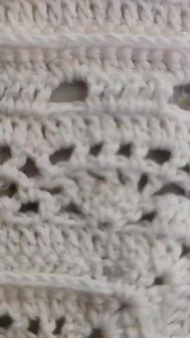 A close-up, static shot of a white crocheted blanket with pink accents at the top. The blanket features various crochet stitches, including openwork patterns, rows of bobble stitches, and solid rows of double crochet. The camera slowly zooms out, revealing more of the blanket's intricate design and the soft color gradient from white to pink at the top. The lighting is soft and even, highlighting the texture of the yarn and the detailed craftsmanship of the crochet work. There are no people or other objects visible in the frame. The overall impression is one of delicate artistry and handmade quality.