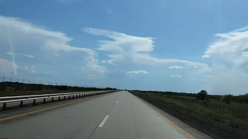 A forward-facing point-of-view video shows a paved highway stretching into the distance under a bright blue sky with scattered white clouds. The highway has two lanes in each direction, separated by a yellow line. A white guardrail runs along the left side of the road, with a strip of vegetation and trees beyond it. The right side of the road is bordered by a grassy field with scattered trees and a barbed wire fence. Several cars are visible on the highway in the distance. The sun is shining, casting a lens flare in the upper left corner of the frame. The weather appears to be clear and fair.