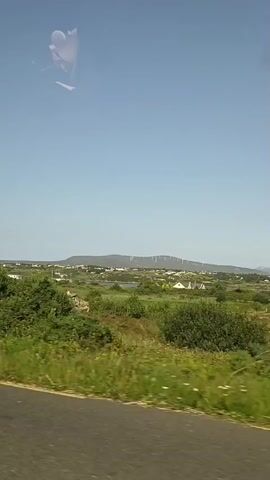 The video begins with a close-up of a white and black camper van. The camera then pans to reveal a rural landscape under a clear blue sky. The scenery includes green fields, trees, and a few houses. One house is made of stone and has a gray tiled roof. Another house is white with a red car parked in the driveway. The video appears to be shot from a moving vehicle.