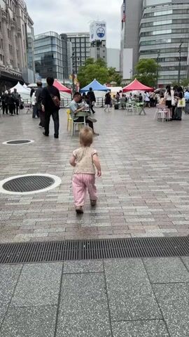 A toddler wearing a light-colored top with dark patterns, pink and white striped pants, and sandals walks on a paved outdoor plaza. The toddler initially walks away from the camera, then turns its head to the left, then turns its body to the right, briefly smiles towards the camera, and then continues walking towards the left side of the frame. The camera follows the toddler at a low angle. In the background, several adults are visible; some are seated at small tables with chairs, while others are walking. Colorful tents are set up further back, suggesting an outdoor market or event. Tall city buildings are visible in the distant background under an overcast sky. The ground is a mix of grey and brown paver stones, with a circular manhole cover and a metal drainage grate visible in the foreground.