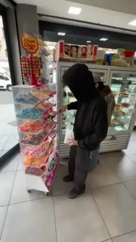 Inside a brightly lit store, a person wearing a black hoodie stands in front of a candy display, filling a white bag with sweets. The display is a multi-tiered, clear plastic container on wheels, stocked with various types of colorful candies. A Chupa Chups lollipop display stands adjacent to the candy rack. To the right is a glass-fronted refrigerator case filled with what appears to be pastries. The floor is tiled in a light grey pattern. The hoodie obscures the person's face. Outside, a street is visible through a glass door.