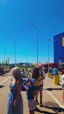 The video captures an outdoor daytime event under a clear blue sky. A paved area with cobblestones is filled with numerous people, including adults and children, walking, standing, and interacting. Tall streetlights are visible, along with multiple flags on poles, some yellow, some blue, and others with colorful floral patterns. In the background, there are trees, white tents, and a stage equipped with speakers. Several individuals are seen bending down, seemingly focused on something on the ground, while others move casually through the scene. Yellow barrels are positioned along the edge of the paved area.