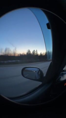A car is driving on a highway at dusk, as seen through a distorted lens mounted inside the vehicle. A line of trees is visible beside the road under a sky tinged with the colors of sunset. The car's side mirror reflects the road and other vehicles, one of which overtakes the car from which the footage is taken. The interior of the vehicle is dark, obscuring details other than the side mirror and part of the window.