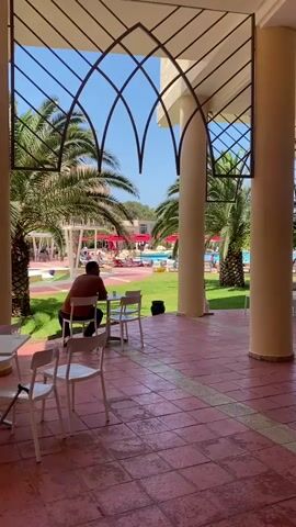 The video shows an outdoor patio area with a man seated at a table, his back to the camera. He is wearing an orange shirt. There are multiple white chairs around the table. The patio has red-tiled floors. Beyond the patio, the scene opens up to a grassy area with several palm trees. Further in the background, a swimming pool surrounded by lounge chairs and red umbrellas is visible. The sky is clear and blue. An ornate metal structure frames the view towards the pool area. The sound of music or instruments can be heard.