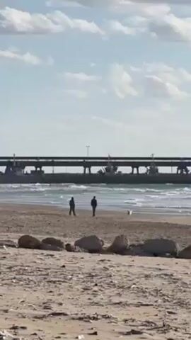 The video shows a beach with two people and a dog walking in the distance. In the background is a pier-like structure with machinery. The sky is blue with scattered clouds, and the foreground features sand and rocks. The scene is captured from a static viewpoint.