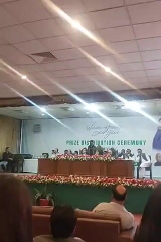 The video shows an indoor gathering, seemingly a prize distribution ceremony. A speaker, wearing a mask, stands at a podium addressing the audience. Behind the speaker, a stage with multiple individuals seated at a table decorated with flowers is visible. A backdrop reads "Prize Distribution Ceremony" and "Welcome to honorable Chief Guest." The audience is seated in rows of chairs, with some people wearing traditional Pakistani clothing. A few people can be seen standing near the stage. Camera equipment is visible on the left. The room is brightly lit by overhead lights.