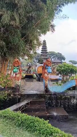 The video shows a scenic view of a temple complex situated on the edge of a body of water. In the foreground, there are lush green plants and bamboo trees. A stone pathway leads towards the temple, flanked by decorative dragon statues with water spouts. These statues are colorful, featuring shades of red, gold, and blue, and appear to be guarding the entrance. There are also intricate blue dragon-like structures extending into the water. Beyond the entrance, a multi-tiered temple structure stands prominently. The water is calm, reflecting the cloudy sky and the surrounding landscape, which includes more temple structures and trees on distant islands or the opposite shore. The overall atmosphere is serene and picturesque.