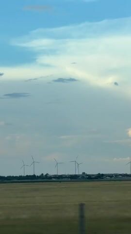 The video displays a wide, horizontal shot of a rural landscape under a cloudy sky. In the foreground, a blur suggests motion, possibly from a vehicle, with a dark fence-like structure visible at the very bottom edge of the frame. The middle ground consists of a field of dry, golden grass or crops. A line of several wind turbines stands in the distance, silhouetted against the sky. Behind the turbines, a low cluster of buildings and trees can be discerned. The sky is a mix of pale blue and white clouds, with a brighter area near the horizon. The overall impression is one of open space and a partly overcast day.