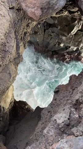The video shows a close-up, overhead view of churning turquoise water inside a rocky cavern. The cavern is formed by dark, volcanic-looking rock. The water is foamy and turbulent. The view is from above, looking down into the opening of the cavern. No people or other objects are visible.