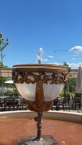 A close-up of an ornate fountain with a clear glass basin and bronze accents. Water sprays from the center of the basin into the air. The fountain is located outdoors, with a bright blue sky and scattered white clouds visible above. In the background, there are tables and chairs set up, suggesting an outdoor seating area for a restaurant or cafe. Umbrellas are visible, and some trees and greenery are in the distance. The overall setting appears to be a pleasant, sunny day.