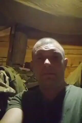 This is a short video clip featuring a man in what appears to be a dimly lit, rustic indoor setting. The walls are made of wooden planks, and there's a canvas or tarp overhead, suggesting a makeshift shelter or a basic room. To the left, there are some piles of what looks like clothing or military gear, camouflaged and in olive green tones. A thick log or post is also visible behind the man. The lighting is dim, creating shadows and highlighting the textures of the wood.

The man is bald and wearing a dark green t-shirt. He is looking directly at the camera and making hand gestures. Initially, he holds up two fingers in a peace sign. Then, he moves his hand down, and it's difficult to discern the exact gesture, but it could be a thumbs down or a similar negative sign. Finally, he makes another hand gesture with his fingers, possibly indicating a shape or a symbol, bringing his hand close to his face.

The man's expression is neutral, and the hand gestures seem to be the main focus of his actions. The overall atmosphere is somewhat raw and unrefined, possibly indicating a temporary living space or a less formal environment. There is no visible text in the video. The context of Zarichne, Ukraine, might suggest a situation related to current events or a rural lifestyle. The time of day is not clearly indicated, but the dim lighting suggests it's likely evening or indoors with limited natural light.