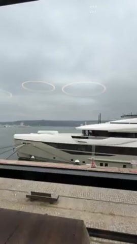 From a slightly elevated, indoor perspective, the video shows an exterior scene featuring a large, modern yacht docked in a harbor. The yacht is primarily white with gray accents. The camera pans right, shifting focus from the yacht to a lighthouse in the distance. The lighthouse is white with a red top and a black base, situated on a small rocky islet. The background consists of a coastline with buildings and trees under an overcast sky. The foreground contains elements of the building the video is shot from, including a window frame, lights, and a bench outside the window.