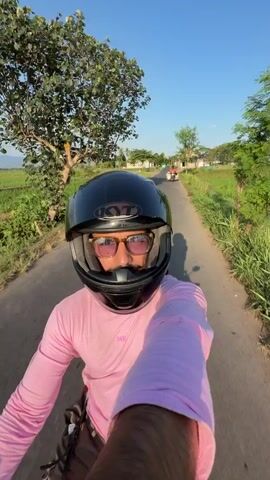 The video begins with a close-up selfie of a person wearing a black helmet and a pink long-sleeved shirt, riding a motorcycle on a paved road. The person is wearing glasses and looking into the camera. The background shows a rural landscape with lush green rice paddies on either side of the road, a tree, and a clear blue sky. As the video progresses, the camera pans to show the expansive rice fields, distant hills, and several coconut trees lining the road. The motorcycle continues to move forward, capturing the scenic views of the countryside under the bright sunlight. There is another person on a motorcycle in the distance, also traveling along the road. The overall scene depicts a peaceful and picturesque journey through a rural area, likely in Southeast Asia given the presence of rice paddies and coconut trees.