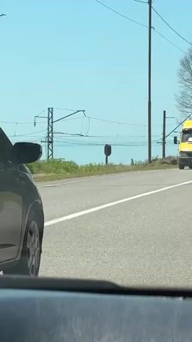 The video captures a sunny daytime drive along a coastal road in Primorskoe, Abkhazia. The scene alternates between external views of the road and an interior shot of a vehicle.

At the beginning, a black hatchback is seen in the foreground, with a yellow van further ahead on the asphalt road. Utility poles and bare trees line the roadside, with a glimpse of a blue body of water (likely the sea) in the distance under a clear sky.

The perspective shifts inside the vehicle, showing a rosary-like beaded ornament hanging from the rearview mirror, swinging slightly. Through the windshield, other cars are visible on the road ahead, and the blue water remains in the background.

The final shot focuses on the road ahead, featuring a white line, green grass, and more trees and utility poles. The blue water stretches to the horizon under the bright sky. Parts of the car's dashboard and a windshield wiper are visible in the foreground, confirming the perspective is from inside a moving vehicle.