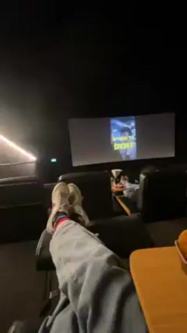 The video starts with a shot of a person's legs, wearing white sneakers and grey pants, with a movie screen in the background, showing a blurry image of a person. There is a small wooden table next to the seat with a basket of food on it. The camera quickly pans to the right to show a young woman seated in the cinema. She gives a thumbs up to the camera, smiling. The cinema is dark, with leather seats.