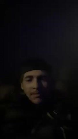 The video features a young man wearing a black beanie and a dark jacket, appearing to be indoors or in a dimly lit environment. His face is partially illuminated, and he looks directly at the camera. There's faint music playing in the background. He makes a peace sign at one point.