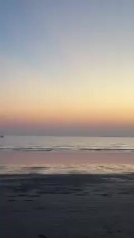 The video shows a beach during sunset. The sky has a gradient of colors, from light blue at the top to orange and pink closer to the horizon. The sea is calm, reflecting the colors of the sky. A small sailboat is visible in the distance, towards the left side of the frame. A person is walking along the wet sand near the water's edge, their silhouette reflected in the water. The beach itself is dark and wet.