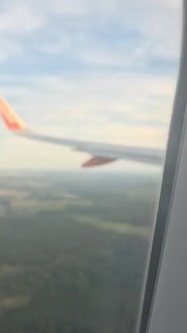 The video is a first-person view from an airplane window, looking out at the wing of the aircraft and the landscape below. The airplane wing is white with an orange tip and the EasyJet logo is visible on the tail of the wing. The landscape below is a mixture of green forests and fields, with some houses and roads visible in the distance. The sky is cloudy with patches of blue. The video appears to be taken during the daytime.