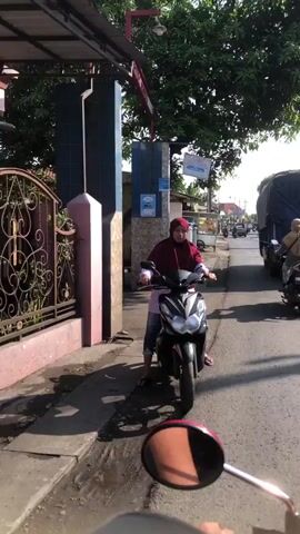 The video shows a person on a motorcycle, wearing a red headscarf and a blue and white shirt, at what appears to be a roadside. The person is looking forward, and the motorcycle is stationary. To the left of the motorcycle is a pink and gray pillar and a decorative metal fence. Behind the pillar is a building with blue siding. The road itself is paved, and there are other vehicles and trees visible in the background. The perspective suggests the video is being filmed from another motorcycle in front of or beside the stationary motorcycle. The lighting indicates it is daytime, possibly morning or afternoon, with sunlight casting shadows.