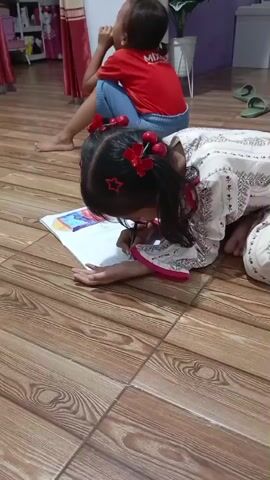 The video opens with a low-angle shot of two children sitting on a wooden floor. The child in the foreground, a young girl with dark hair adorned with red hair clips, is on her knees, intently focused on a drawing in front of her. She wears a white and pink patterned outfit. Her arms are resting on the floor as she draws. In the background, another child, wearing a red shirt and blue pants, is seated with their back to the camera, their head tilted slightly upwards as if looking at something or someone off-screen. The environment appears to be an indoor space, possibly a living room or play area, with a tiled floor and a glimpse of furniture and curtains in the background. The overall atmosphere is one of quiet activity and focus.