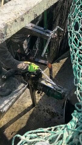 A person wearing a gray long-sleeved shirt, dark pants, and lime green work gloves sprays water on what appears to be the dirty mechanics of a boat engine or motor. The engine is mounted on a metal frame within the confines of a structure covered in what looks like grime. The person stands on a metal footplate, and a green net hangs in the corner of the frame. The sky is blue, and some buildings and trees can be seen in the background.