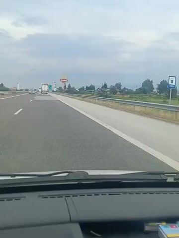 This media file shows a first-person perspective video of driving on a highway. The scene is a multi-lane highway with other vehicles, including trucks and cars, traveling in the same direction. On the right side of the highway, there are signs for McDonald's and a gas station, suggesting a roadside service area. Trees and vegetation line the side of the road, with distant hills or mountains visible in the background. The sky is overcast with clouds, indicating a potentially cloudy or approaching rain day. The time of day appears to be daytime, possibly late morning or afternoon. The overall mood is neutral, conveying a sense of travel. There is visible text on the signs, including "McDonald's" in English. There are no people clearly visible in the frame, and the focus is on the road and the surrounding environment from the driver's viewpoint. The setting is a highway in Gelemenovo, Bulgaria.
