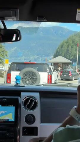 A person in the driver's seat of a vehicle is recording from inside as other vehicles line up at a ferry terminal. A view of a mountain and lake are visible through the front windshield of the car. Other cars and people are passing through the terminal.