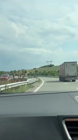 The video shows a view from inside a car traveling on a highway. The car is in the right lane, and a large white truck with a red stripe is in the left lane. The highway has multiple lanes and is bordered by a metal guardrail and grassy areas. In the background, there are rolling hills with trees and vegetation. Power lines and transmission towers are visible on the hills. The sky is partly cloudy with some darker clouds suggesting a possibility of rain. The overall scene depicts highway travel in a rural or semi-rural landscape.
