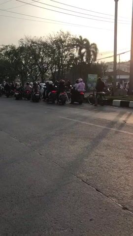 The video shows a street scene with a line of motorcycles and their riders. The motorcycles are parked or moving slowly in a line, suggesting a traffic jam or a gathering. The riders are mostly wearing helmets. Some motorcycles have passengers. In the background, there are trees, buildings, and utility poles. The sky is visible, with sunlight suggesting it is either early morning or late afternoon. A bicycle is visible to the right of the frame. The road appears to be paved.