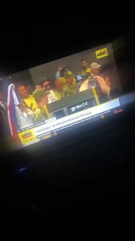 The video shows a laptop screen displaying news footage. The news report features a crowd of people surrounding a figure, possibly Ronaldinho, taking photos and reaching out to him. The crowd includes individuals wearing yellow shirts, military uniforms, and various hats. The news graphic at the bottom of the screen mentions "GESTORES" and "CASO RONALDINHO". The laptop keyboard is visible in the foreground, suggesting the video is being recorded from a personal device.