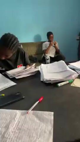 Three young people are in a room with light blue walls. Two of them are seated at a table piled high with papers, pens, and a phone. The third person sits on a couch in the background. The two at the table appear to be working or studying with the papers, while the person on the couch seems to be resting. One of the people at the table picks up sheets of paper and looks at them. The other person is looking at a phone. A doorway is visible on the other side of the room.