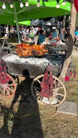 A whole pig is roasting on a spit over coals in a rustic metal cart. The cart has two large wooden spoked wheels with red and black checkered cloths hanging from them. In the background, there are people browsing at an outdoor market, tents, and green umbrellas. A Bulgarian flag is visible on the right.