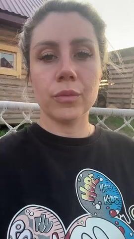 A woman with blonde hair pulled back in a bun is wearing a black t-shirt with a Mickey Mouse graphic. She is holding a silver can to her lips and drinking. She is sitting outdoors in front of a wooden fence, with a hammock visible behind her. The sun appears to be setting, casting a warm light on the scene.