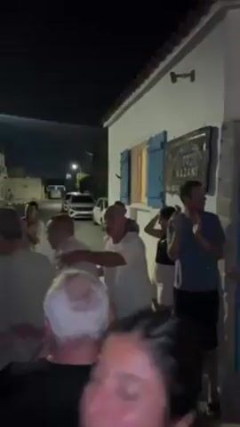 The video captures a lively scene at night where a group of about 20 people, a mix of adults and young adults, are dancing in a street. They appear to be celebrating or enjoying a spontaneous gathering, as they move enthusiastically to music. Most are wearing casual clothing, light-colored shirts are prominent. The setting is a narrow street between white buildings, typical of a Mediterranean town. A sign in Greek reads "TO KAZANI" on one of the buildings. A street light illuminates the dancing group, casting shadows and enhancing the festive atmosphere. The overall vibe suggests a joyful and communal experience.
