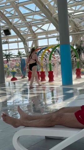 The scene unfolds inside an indoor water park, characterized by a sprawling, glass-paneled ceiling supported by a geometric network of white metal beams. In the foreground, a man lies on a white plastic lounge chair, shirtless and clad in red swimming trunks, with his feet pointing towards the camera. He appears relaxed, with his eyes closed and arms folded across his chest.
Beyond the man, a woman in a black swimsuit is seen walking away from the camera through an area with spraying water features. These colorful structures, resembling a playful water playground, include archways, spouts, and whimsical, tree-like features that spray water. The floor is covered in a shallow layer of water, reflecting the overhead light and adding to the aquatic ambiance. Further back, faux palm trees add a touch of tropical decor to the indoor water park setting.