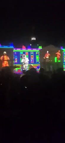 The video shows a nighttime outdoor projection mapping show on the facade of a building that resembles a palace or grand house. The projection displays stylized figures, some appearing to be in orange protective suits and others in what look like hazmat suits or similar attire. The projections are highly colorful, with vibrant greens, blues, pinks, and oranges. The figures are arranged in a way that suggests a performance or art installation. In the foreground, silhouetted by the projection light, are the heads and shoulders of a crowd of people watching the show. The overall atmosphere is dark, with the building and its projections being the primary illuminated elements. The camera is handheld and the footage is slightly shaky.
