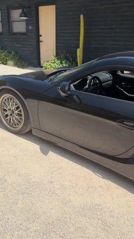 The video shows two sports cars parked outside a building. The first car, a black sports car, is parked in the foreground on the left side of the frame. The second car, a silver Toyota Supra, is parked in the background, facing away from the camera. A silver Porsche convertible is parked behind the Supra. The building appears to be made of dark wood or brick and has a light-colored door. There are some plants and cacti around the building. The ground is covered in gravel or dirt. The video pans slightly to the left and then to the right, showing different angles of the cars and the environment.