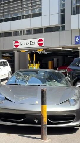 A grey Ferrari 458 Italia is parked in the foreground, partially obscured by a black and yellow bollard. Behind it, a white SUV is visible on the left. In the background, a red car, possibly a Porsche Macan, is slowly exiting a parking garage. Above the garage exit, a sign reads "Izlaz-Exit" with two red "do not enter" symbols. A man wearing a light-colored shirt and trousers walks from left to right behind the white SUV. Another sign reading "Naplata Parkinga" is visible to the left of the garage entrance. The scene appears to be an outdoor parking area on a bright day.