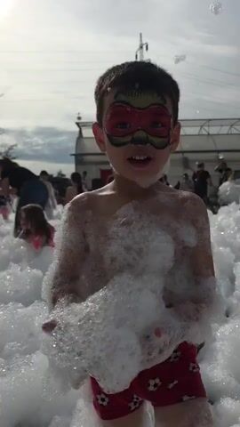 The video shows a child's eye view of a foam party. The child, wearing a red mask face paint, is holding a bunch of foam in front of the camera, obscuring the view.  The camera then pans out, revealing numerous children playing in a large pile of white foam. Several adults are visible amongst the children, presumably supervising. A white building, possibly a greenhouse, and power lines are visible in the background, suggesting the party is taking place outdoors. The child with the face paint looks up at the camera and smiles, their face covered in foam.