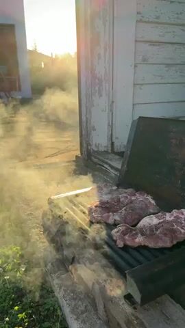 The video shows a close-up, low-angle shot of raw steaks grilling on an outdoor barbecue. The steaks are pink and appear to be seasoned with salt and pepper. Smoke rises from the grill, obscuring some of the meat. The grill itself is made of black metal and rests on rough-hewn wooden supports. To the right of the grill is the side of a weathered, white wooden building with peeling paint. Sunlight streams in from the left, creating a lens flare and illuminating the smoke. The background shows a paved area, some greenery, and the edge of another white building. The overall impression is one of an outdoor cooking scene at sunset.