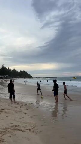 A group of young males are playing soccer on a wet sand beach. The sky is overcast with dramatic clouds. Several other people are visible in the background, along the shoreline and in the ocean. The waves are moderate, and there appears to be a small yellow watercraft in the distance. The overall scene is one of casual recreation on a beach.