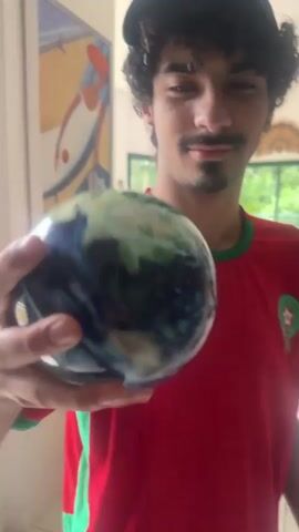 Here is a detailed description of the video:

A young man stands indoors, holding a globe in his right hand. He is wearing a red soccer jersey with green trim, a dark baseball cap, and has curly hair and a mustache. The jersey has the Puma logo and the crest of Morocco. He looks directly at the camera, holding the globe up in front of his face. 

The background reveals an interior space with tiled flooring and white walls. Behind him, there is artwork hanging on the wall, and a glimpse of a room with a large window showing greenery outside.  An orange chair and other furniture are visible. There is also an audible ruckus happening in the background while he poses with the globe.