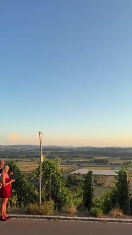A vertical, handheld video shows a scenic landscape at sunset. The sky transitions from a clear blue at the top to a warm orange and yellow gradient near the horizon. In the foreground, lush green grapevines are planted along a slope, with wooden posts supporting the vines. A paved road is visible at the bottom of the frame. Beyond the vines, a patchwork of agricultural fields stretches out, dotted with a few buildings. As the video progresses, several cars are parked along the roadside, suggesting a gathering of onlookers. The sun, a bright, intensely lit orb, is positioned lower in the sky, casting a golden glow over the scene and creating lens flares. The sun's appearance gradually changes, indicating a solar eclipse is taking place, with a crescent shape becoming more pronounced as the video nears its end. The overall atmosphere is serene and picturesque, capturing a natural phenomenon against a beautiful backdrop.
