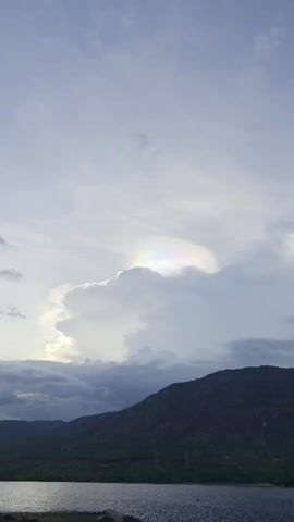The video shows a scenic view with a body of water, mountains, and a cloudy sky. The camera zooms in on a specific cloud formation displaying a colorful iridescence or a partial rainbow effect along its edge. The sky is overcast, and the mountains are dark green with some visible structures or power lines. The water is calm with some ripples. The overall lighting suggests either early morning or late afternoon.