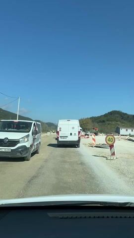 The video shows a view from inside a car driving on a road that appears to be under construction. The road surface is uneven and dirty, with areas of exposed gravel and dirt. There are several white vans in front of the car, driving in the same direction. Construction equipment and materials are visible on the sides of the road, including red and white striped barriers and construction trailers. A speed limit sign indicating 40 is visible on the right side of the road. Hills with green vegetation can be seen in the background, under a clear blue sky.
