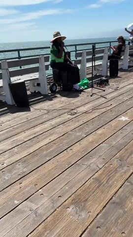 A person wearing a straw hat and a green shirt sits on a trolley next to a speaker, singing into a microphone. Another person sits on a bench to the right, also with a speaker. The pier is made of wooden planks. In the background, the ocean and a blue sky with clouds are visible. Other people are seen further down the pier, some holding umbrellas. The sun is shining.