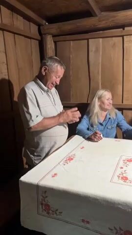 The video shows an indoor scene with wooden walls and a wooden ceiling with beams. A group of people are gathered around a table covered with a white tablecloth featuring a floral pattern. On the left, an older man in a light-colored short-sleeved shirt is clapping his hands and smiling. To his right, a blonde woman in a denim jacket is seated at the table, also smiling and holding something in her hands. Next to her, another woman in a blue and white jacket is seated and looking forward, with her hand raised. In the foreground, partially obscuring the view of the table, a person in gray pants is visible. The overall atmosphere appears to be casual and social.