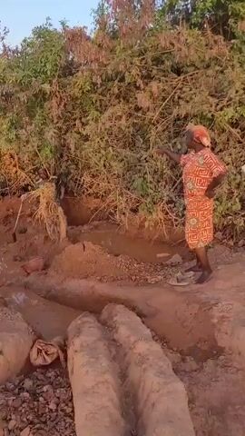 The video shows a muddy area with people working. A woman in an orange dress points to something behind a bush. The camera pans to show a wider view of the area, revealing more people, some with tools, working in a field-like environment. There's a building in the background and individuals walking with items on their heads. The area appears to be under construction or undergoing some sort of earthmoving activity.