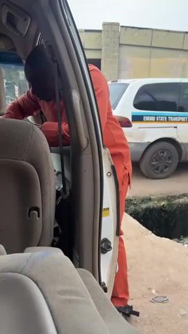 A person wearing an orange jumpsuit is seen leaning into the open passenger door of a white vehicle. The person appears to be interacting with something inside the car. The interior of the car shows beige-colored seats and door panels. Outside the car, a white van with blue and yellow stripes and text reading "ENUGU STATE TRANS..." is visible. The ground appears to be dirt and gravel.