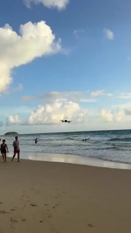A drone is flying over the ocean near a beach. Several people are visible on the beach and in the water. The sky is blue with scattered clouds. The waves are moderate. The sand is light brown.